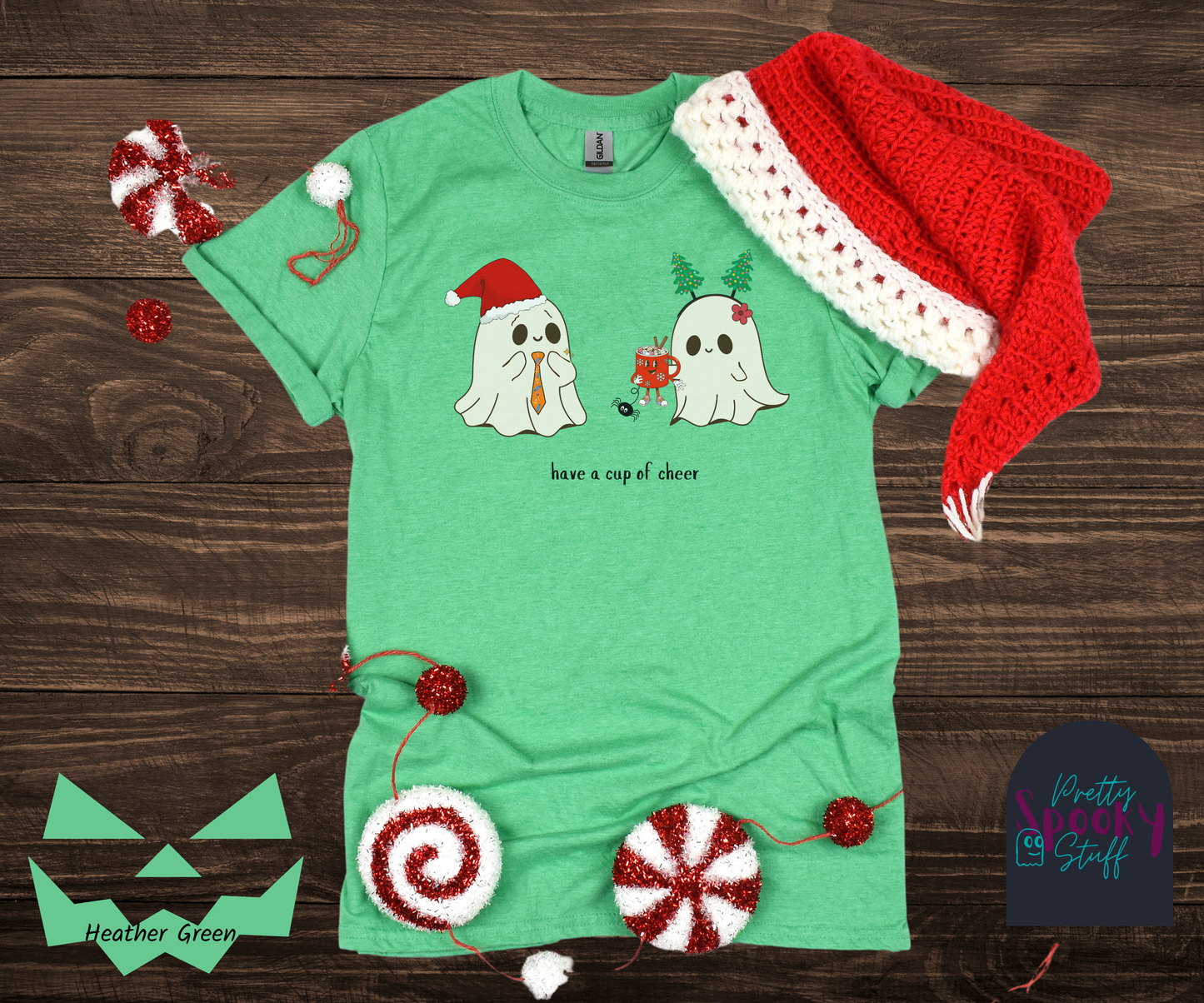 Christmas shirt with two ghosts, one wearing a Santa hat with Halloween candy tie, and one wearing a Christmas tree headband that is handing the other ghost a cup of hot chocolate that has a face and legs, and also a spider hanging from the bottom, with the words "have a cup of cheer" below, in heather green.