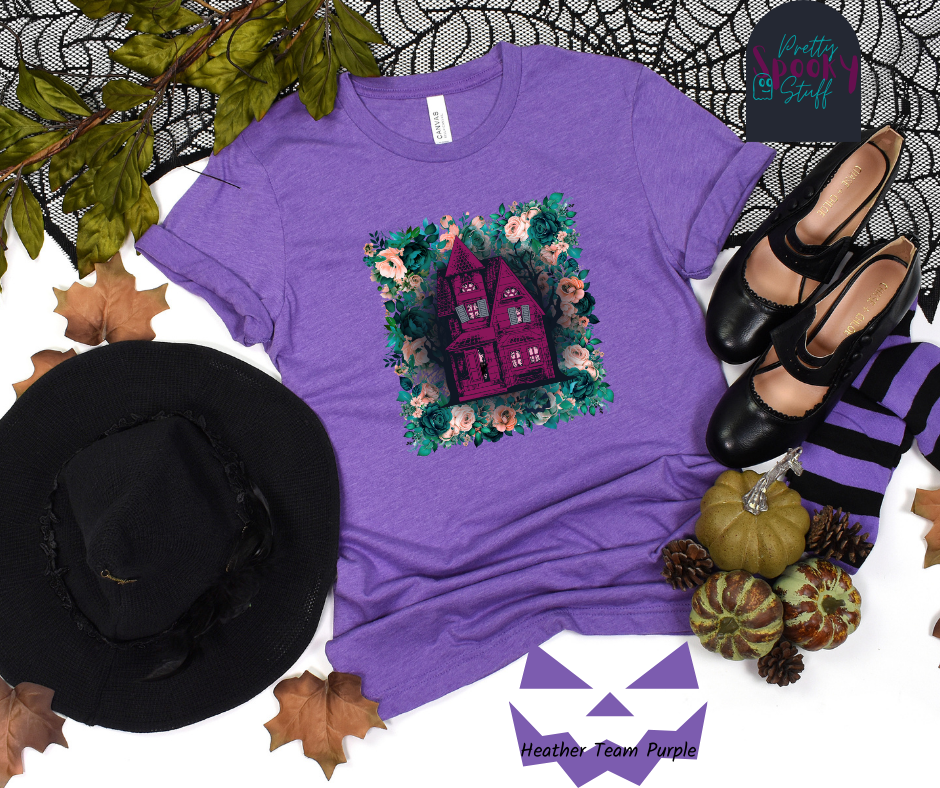 Haunted house Halloween shirt with purple haunted house sitting on top of a rectangle bed of blue and pink flowers, in heather team purple.