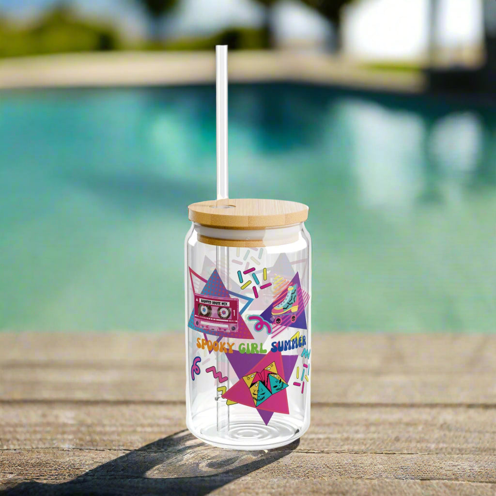 Summerween Glass Tumbler featuring retro 90's design including cassette tape with "haunted house mix", bat covered roller skate, folded paper fortune teller, bright-colored squiggles, and words "spooky girl summer" across the middle in orange, green, and dark blue. Poolside.