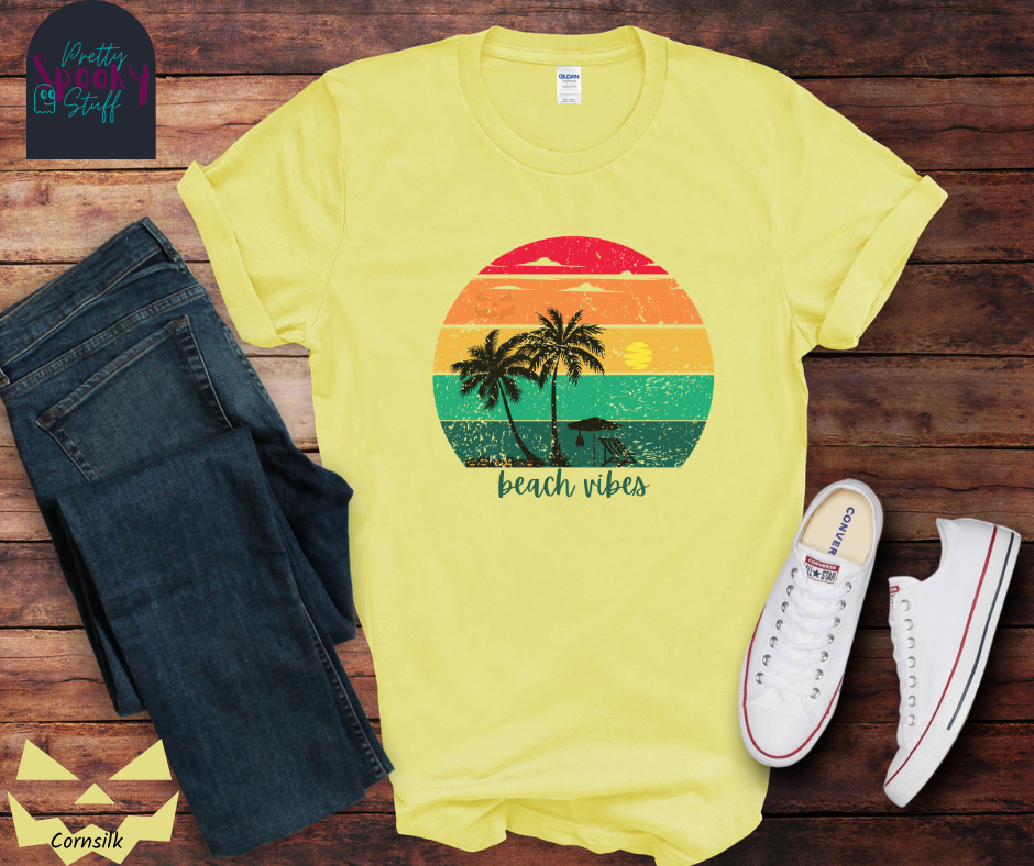 Summerween shirt with rainbow sunset ombre lines, behind the silhouettes of two palm trees, beach umbrella and chair, featuring a bat hanging from the umbrella, and group of jack o'lanterns, with blue calligraphy font in the words "beach vibes", vintage look, in cornsilk.