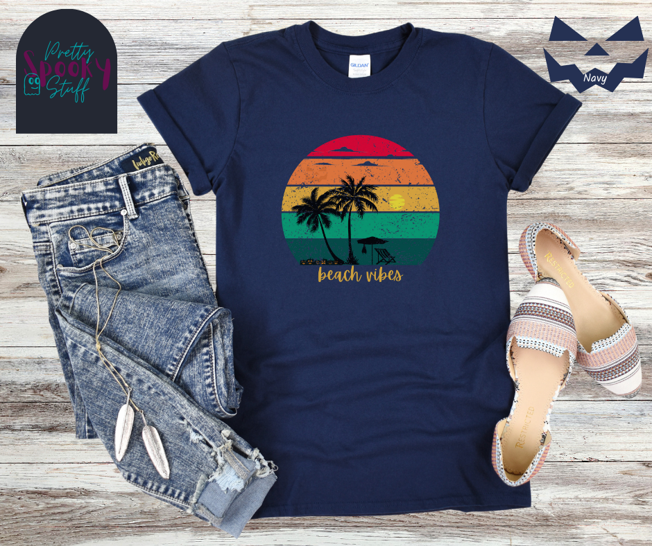 Summerween shirt with rainbow sunset ombre lines, behind the silhouettes of two palm trees, beach umbrella and chair, featuring a bat hanging from the umbrella, and group of jack o'lanterns, with yellow calligraphy font in the words "beach vibes", vintage look, in navy.