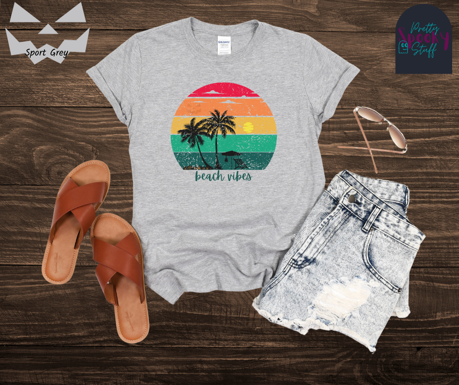 Summerween shirt with rainbow sunset ombre lines, behind the silhouettes of two palm trees, beach umbrella and chair, featuring a bat hanging from the umbrella, and group of jack o'lanterns, with blue calligraphy font in the words "beach vibes", vintage look, in sport grey.