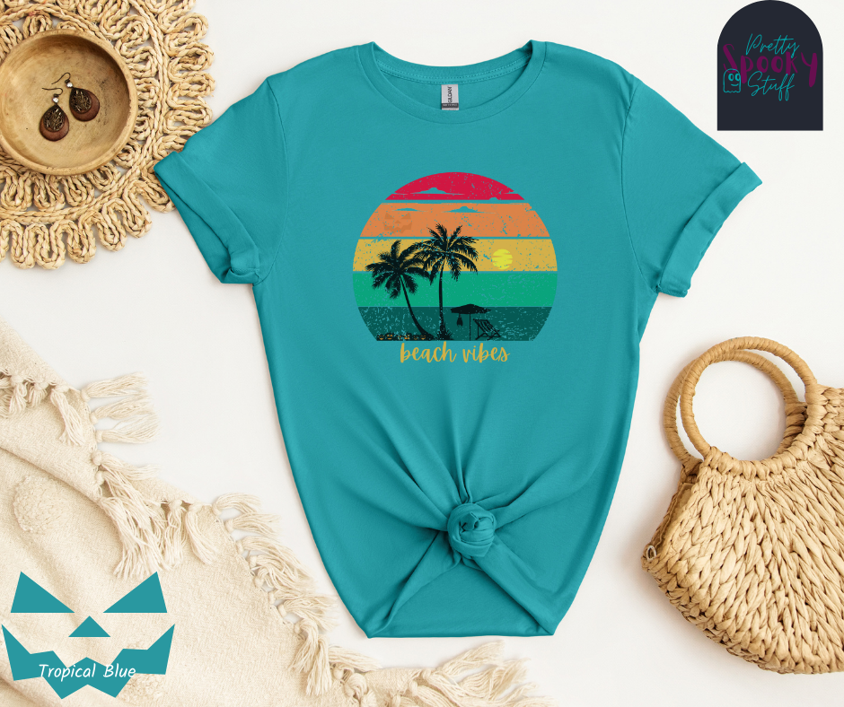 Summerween shirt with rainbow sunset ombre lines, behind the silhouettes of two palm trees, beach umbrella and chair, featuring a bat hanging from the umbrella, and group of jack o'lanterns, with yellow calligraphy font in the words "beach vibes", vintage look, in tropical blue.