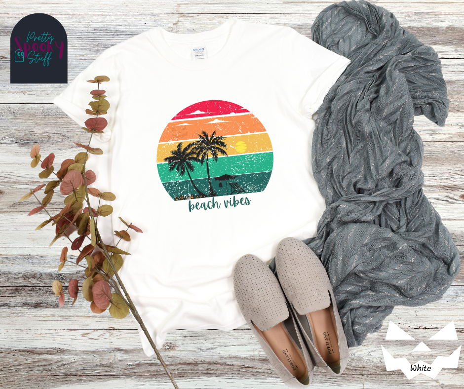 Summerween shirt with rainbow sunset ombre lines, behind the silhouettes of two palm trees, beach umbrella and chair, featuring a bat hanging from the umbrella, and group of jack o'lanterns, with blue calligraphy font in the words "beach vibes",vintage look, in white.