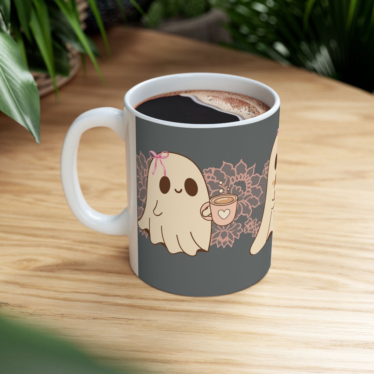 Ceramic Halloween coffee mug with three adorable ghosts, each enjoying their own coffee beverage, adorned with a pink bow, with light pink intricate lace design behind them, gray background, side view sitting on table.