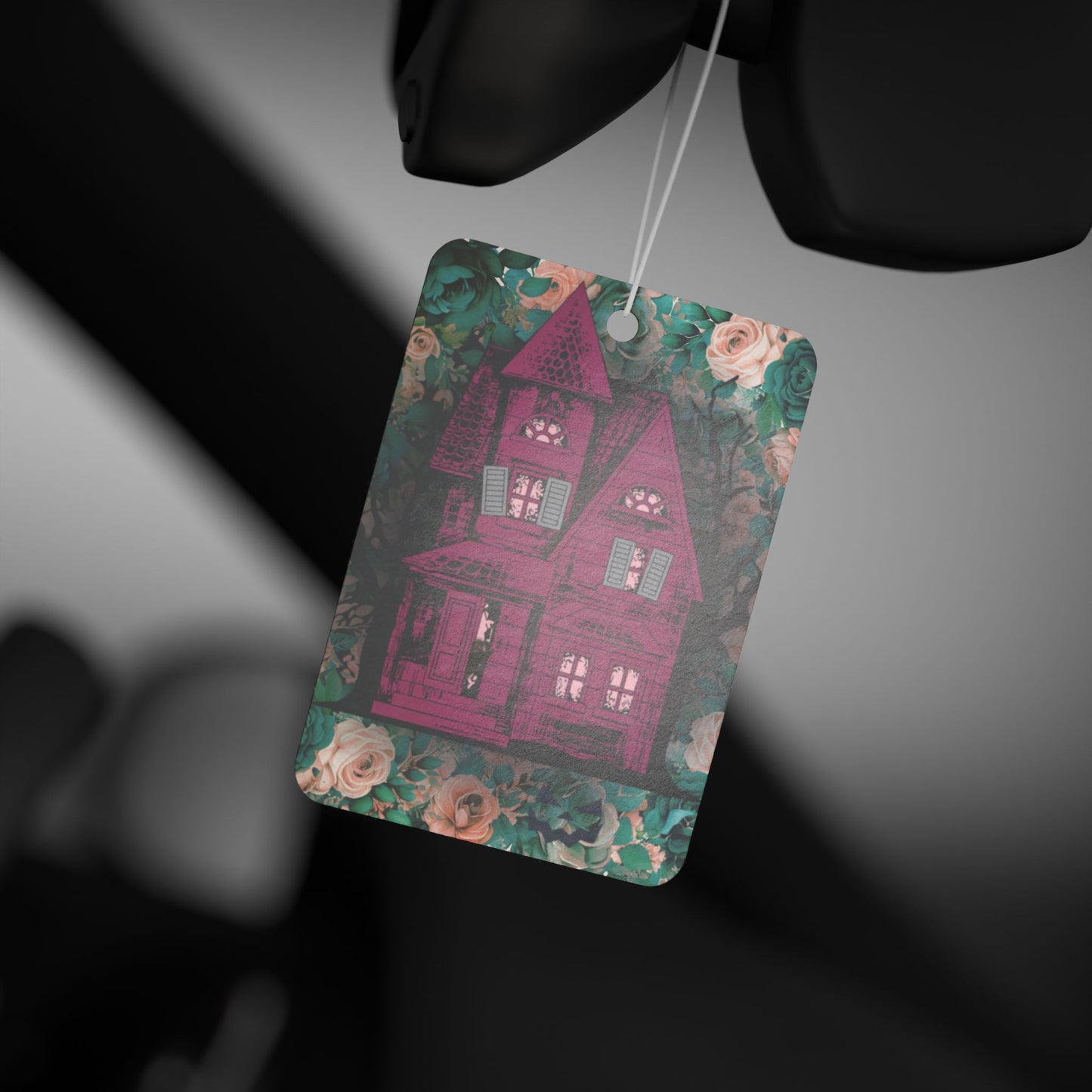 Haunted house Halloween air freshener with purple haunted house sitting on top of a rectangle bed of blue and pink flowers, hanging sideways in car.
