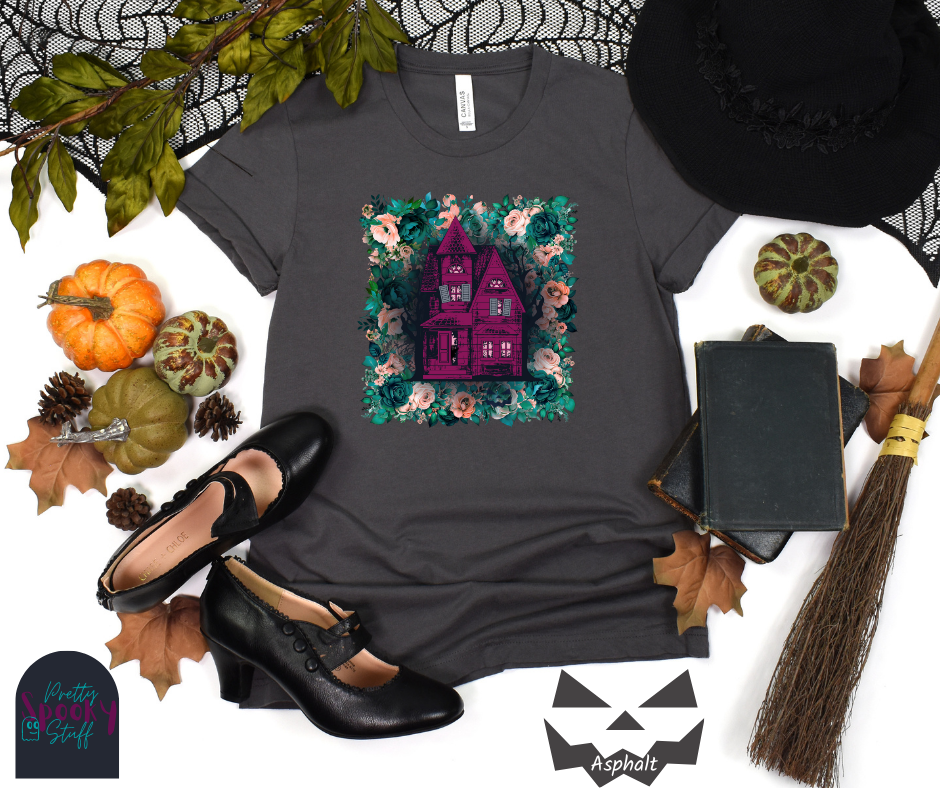 Haunted house Halloween shirt with purple haunted house sitting on top of a rectangle bed of blue and pink flowers, in asphalt.