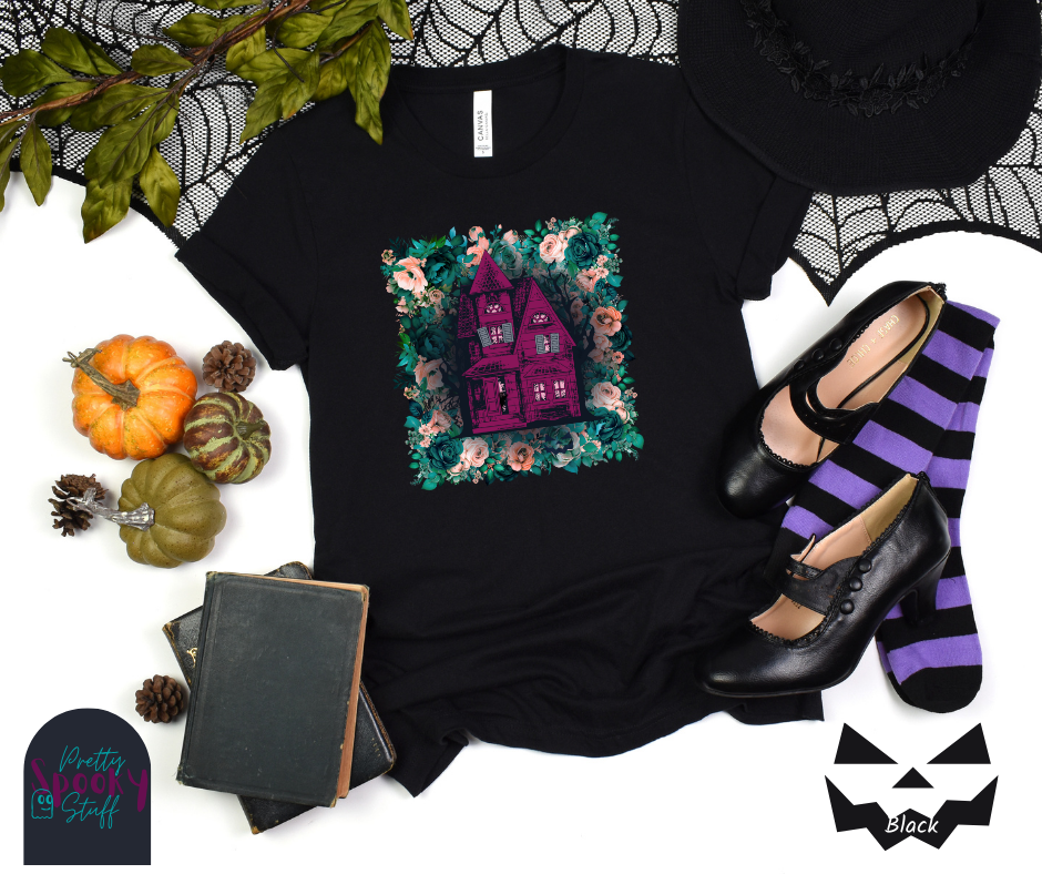 Haunted house Halloween shirt with purple haunted house sitting on top of a rectangle bed of blue and pink flowers, in black.