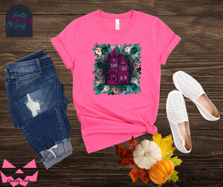 Haunted house Halloween shirt with purple haunted house sitting on top of a rectangle bed of blue and pink flowers, in charity pink.