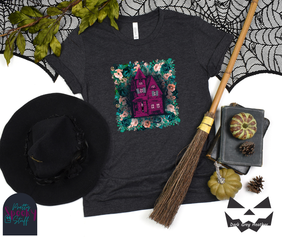 Haunted house Halloween shirt with purple haunted house sitting on top of a rectangle bed of blue and pink flowers, in dark grey heather.