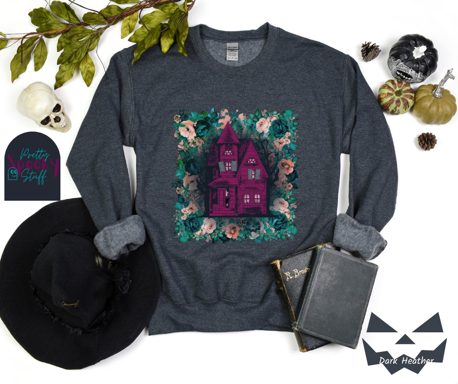 Haunted house Halloween sweatshirt with purple haunted house sitting on top of a rectangle bed of blue and pink flowers, in dark heather.