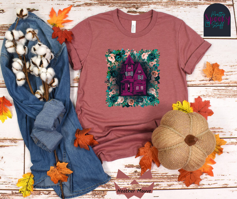 Haunted house Halloween shirt with purple haunted house sitting on top of a rectangle bed of blue and pink flowers, in heather mauve.
