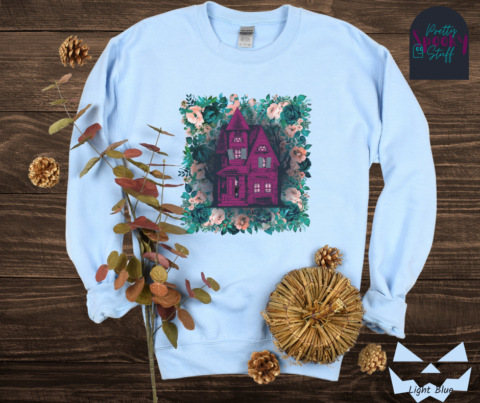 Haunted house Halloween sweatshirt with purple haunted house sitting on top of a rectangle bed of blue and pink flowers, in light blue.