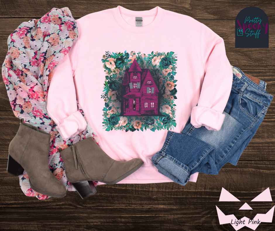 Haunted house Halloween sweatshirt with purple haunted house sitting on top of a rectangle bed of blue and pink flowers, in light pink.