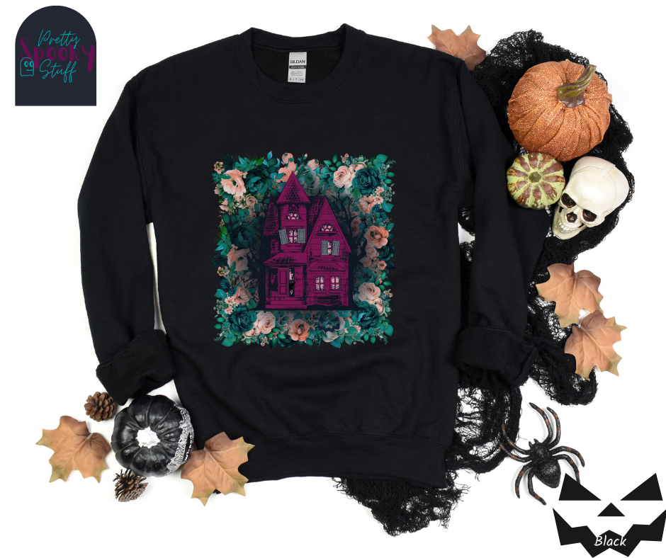 Haunted house Halloween sweatshirt with purple haunted house sitting on top of a rectangle bed of blue and pink flowers, in black.