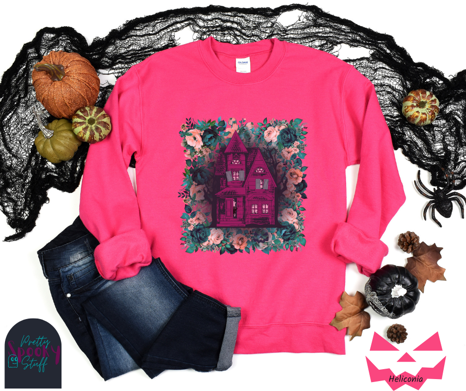 Haunted house Halloween sweatshirt with purple haunted house sitting on top of a rectangle bed of blue and pink flowers, in heliconia.