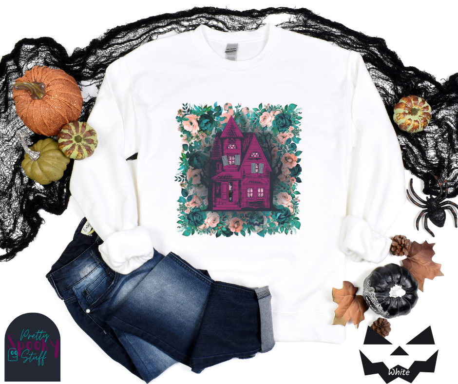 Haunted house Halloween sweatshirt with purple haunted house sitting on top of a rectangle bed of blue and pink flowers, in white.
