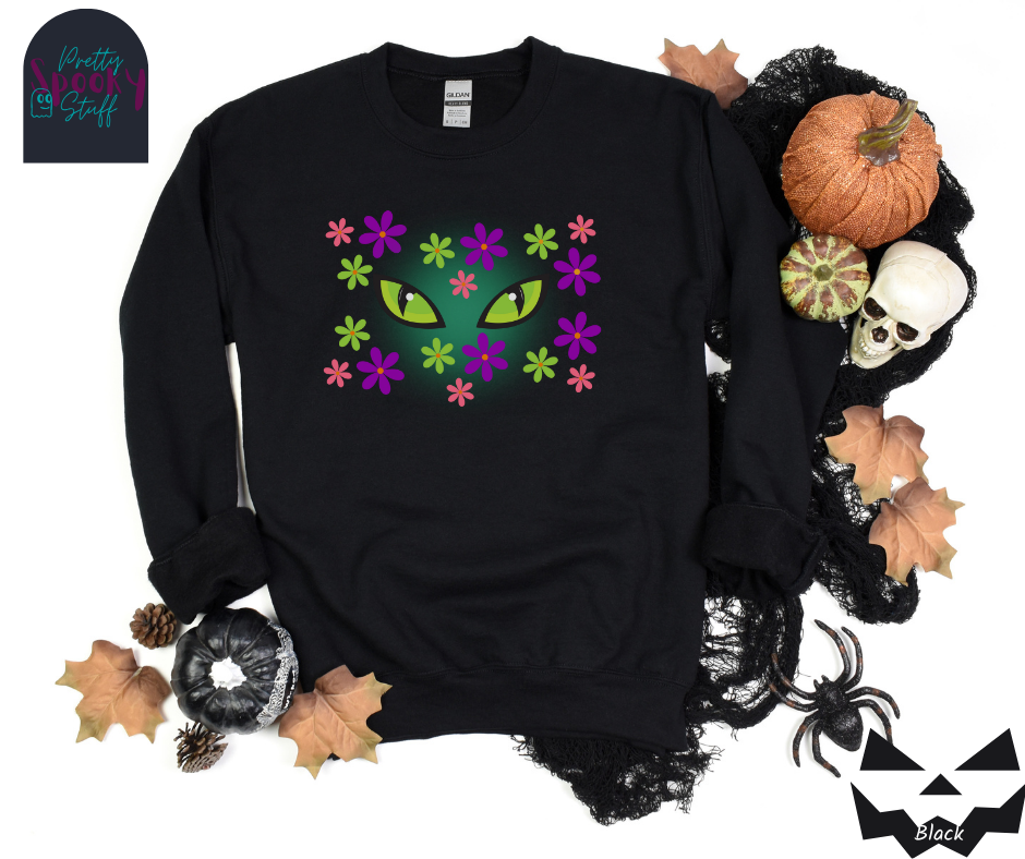 Cat Halloween sweatshirt with spooky green cat eyes in the middle of purple, pink and green flowers, with a blue glow behind them, in black.