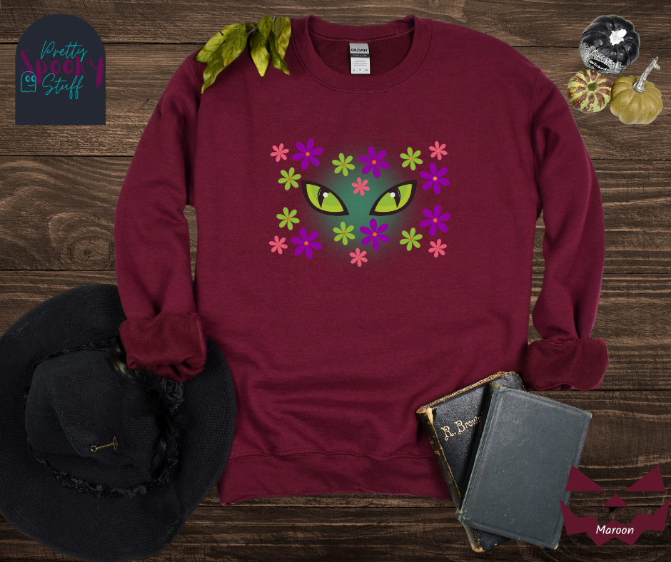 Cat Halloween sweatshirt with spooky green cat eyes in the middle of purple, pink and green flowers, with a blue glow behind them, in maroon.