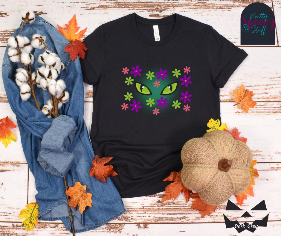 Black cat Halloween shirt with bright green cat eyes in the midst of pink, green, and purple flowers with a blue glow behind them, in dark grey.