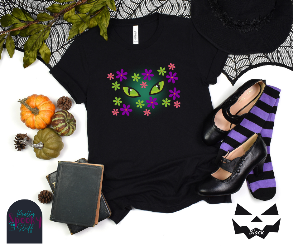 Black cat Halloween shirt with bright green cat eyes in the midst of pink, green, and purple flowers with a blue glow behind them, in black.