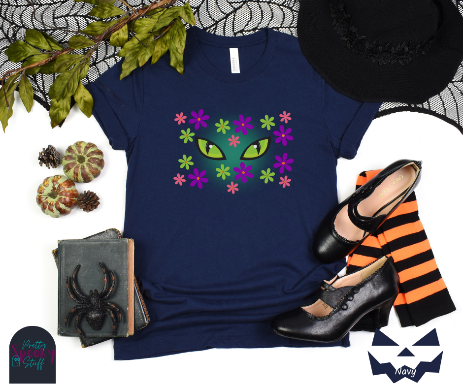 Black cat Halloween shirt with bright green cat eyes in the midst of pink, green, and purple flowers with a blue glow behind them, in navy.