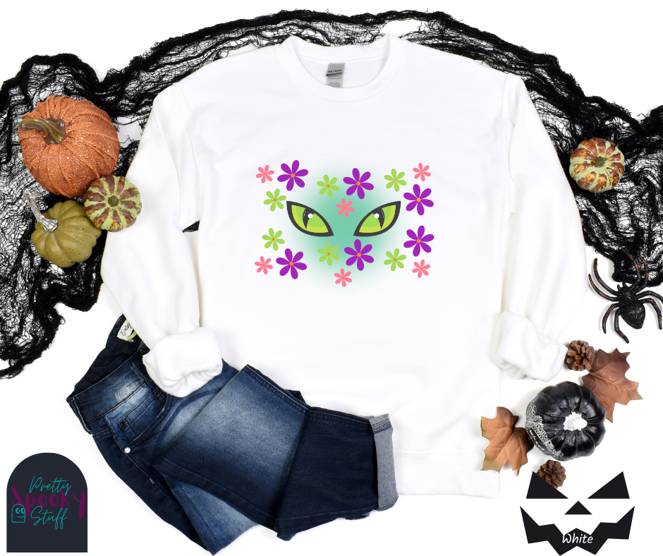 Cat Halloween sweatshirt with spooky green cat eyes in the middle of purple, pink and green flowers, with a blue glow behind them, in white.