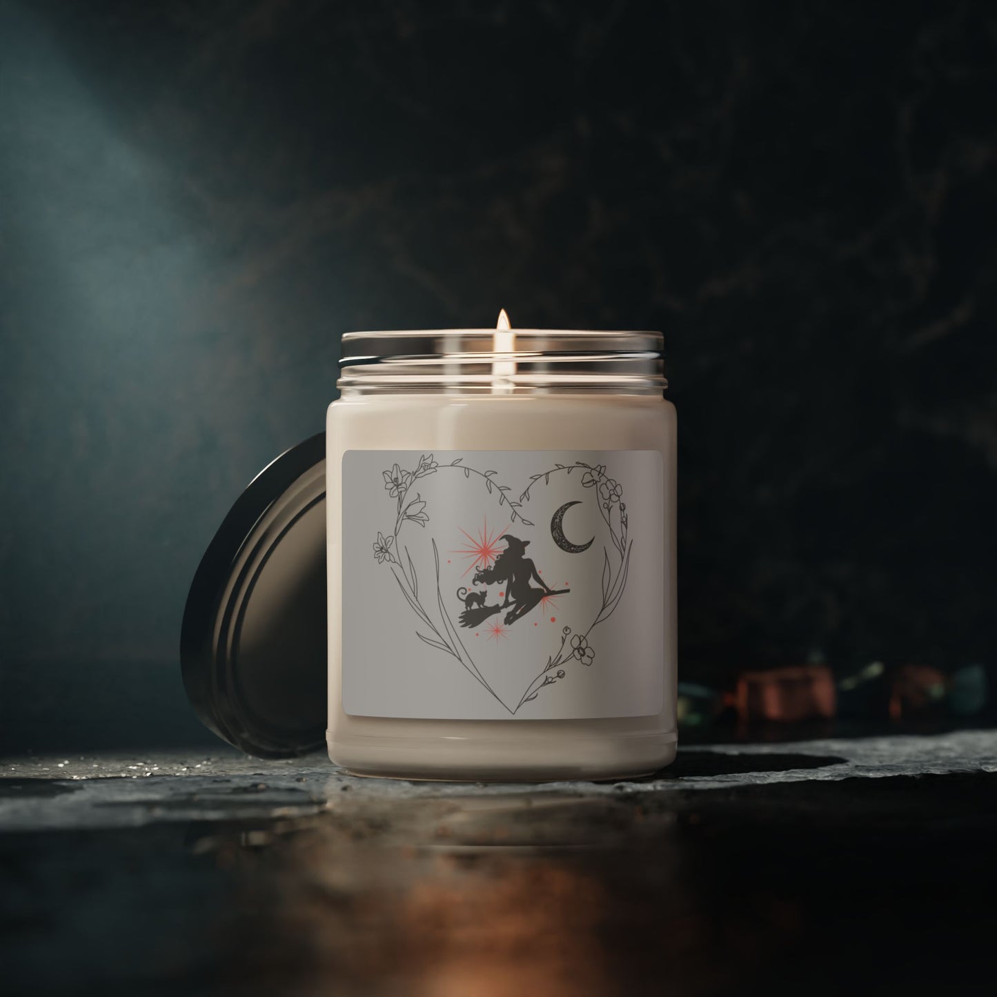 Witch themed soy candle with a silhouette of an elegant with and cat sitting on a broom with pink starbursts behind them, a floral crescent moon, surrounded by a heart formed by delicate stemmed flowers, with light gray sticker background, white wax, in glass jar, sitting on dark table with dark marble wall.