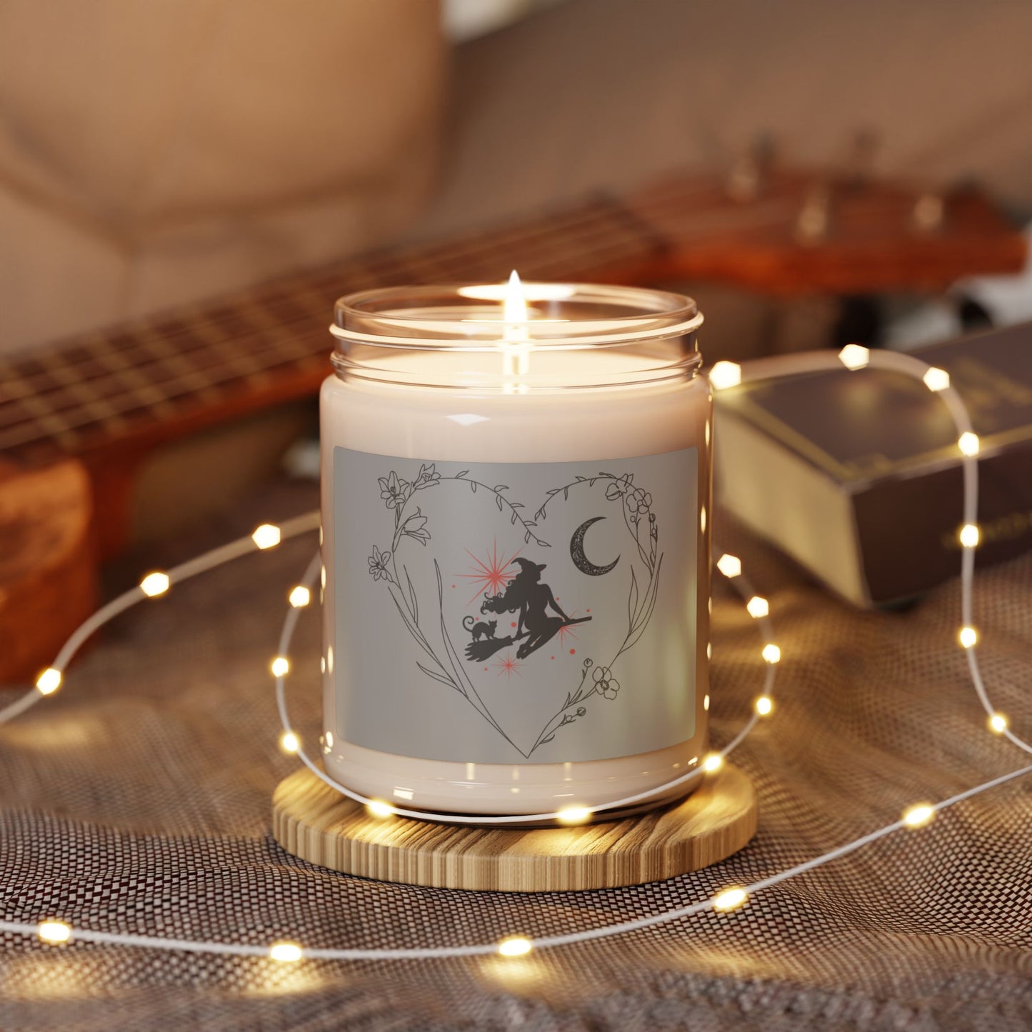 Witch themed soy candle with a silhouette of an elegant with and cat sitting on a broom with pink starbursts behind them, a floral crescent moon, surrounded by a heart formed by delicate stemmed flowers, with light gray sticker background, white wax, in glass jar, sitting on table with guitar and fairy lights.
