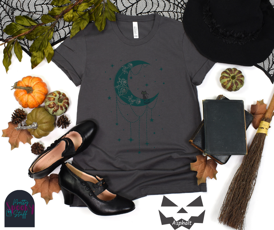 Moon and stars Halloween shirt with a teal crescent moon with pink floral design inside of it, beads hanging from the bottom, a black cat perched on the bottom, and teal stars surrounding it, in asphalt.