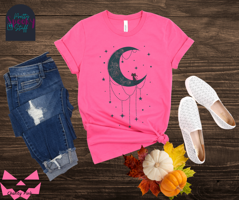 Moon and stars Halloween shirt with a teal crescent moon with pink floral design inside of it, beads hanging from the bottom, a black cat perched on the bottom, and teal stars surrounding it, in charity pink.