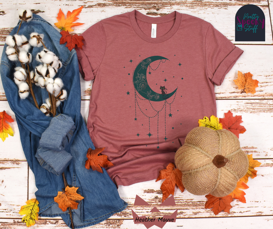 Moon and stars Halloween shirt with a teal crescent moon with pink floral design inside of it, beads hanging from the bottom, a black cat perched on the bottom, and teal stars surrounding it, in heather mauve.
