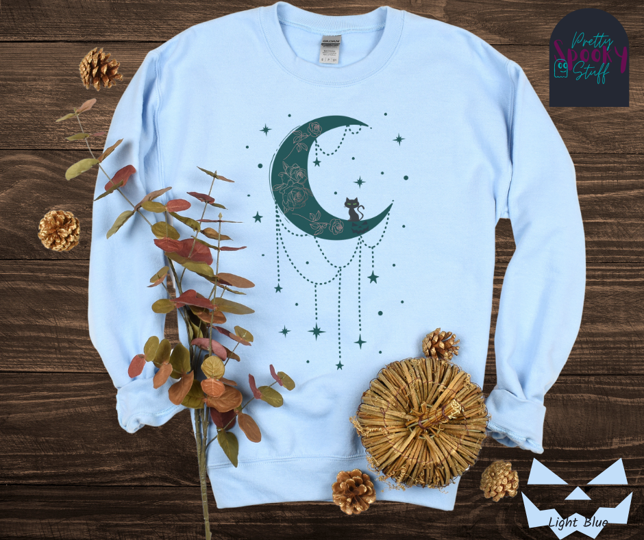 Moon and stars Halloween sweatshirt with a teal crescent moon with pink floral design inside of it, beads hanging from the bottom, a black cat perched on the bottom, and teal stars surrounding it, in light blue.