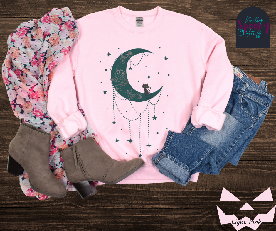 Moon and stars Halloween sweatshirt with a teal crescent moon with pink floral design inside of it, beads hanging from the bottom, a black cat perched on the bottom, and teal stars surrounding it, in light pink.