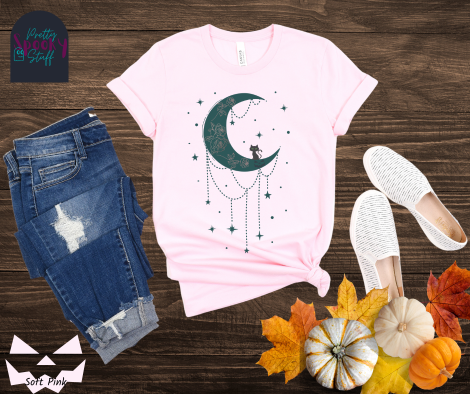Moon and stars Halloween shirt with a teal crescent moon with pink floral design inside of it, beads hanging from the bottom, a black cat perched on the bottom, and teal stars surrounding it, in soft pink.
