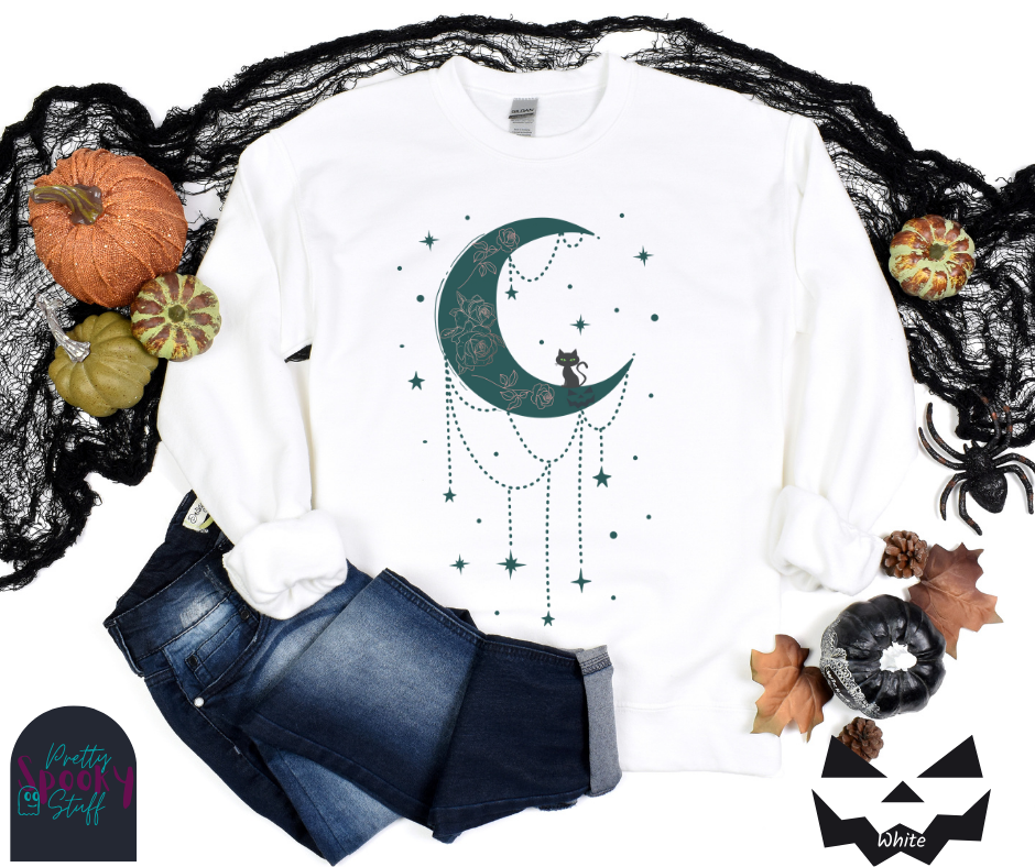 Moon and stars Halloween sweatshirt with a teal crescent moon with pink floral design inside of it, beads hanging from the bottom, a black cat perched on the bottom, and teal stars surrounding it, in white.