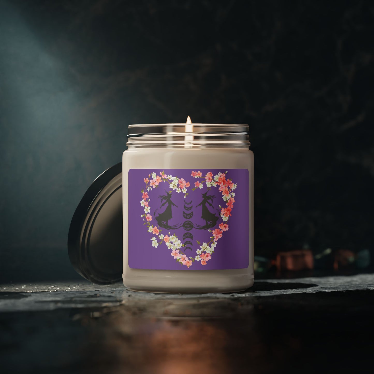 Witch themed summerween soy candle featuring the silhouettes of two graceful witches facing each other in mid glide, the phases of the moon between them with floral details, surrounded by a heart made from pink and white cherry blossoms, with a medium purple sticker background, sitting on a dark table with dark marble wall.