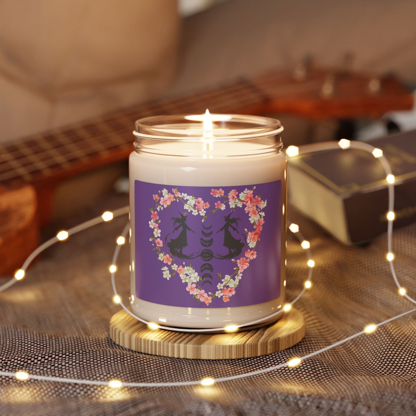 Witch themed summerween soy candle featuring the silhouettes of two graceful witches facing each other in mid glide, the phases of the moon between them with floral details, surrounded by a heart made from pink and white cherry blossoms, with a medium purple sticker background, sitting on a table with a guitar and fairy lights.