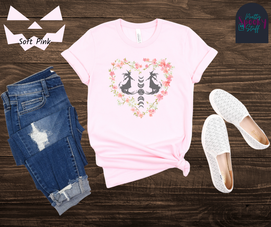 Witch themed summerween shirt with silhouettes of two graceful witches mid-step on what seems like fog, with the moon phases going top to bottom in between them, surrounded by a heart of pink and white cherry blossoms, in soft pink.