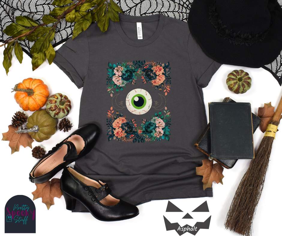 Unique Halloween shirt with large eye in the middle, with gold line drawing of crescent moon and star, and four bunches of pink and blue flowers forming a frame around it, in asphalt.