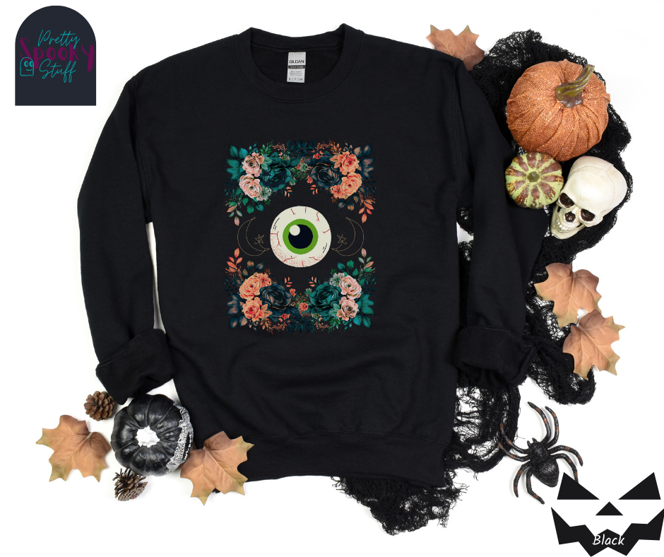 Floral Halloween sweatshirt with a large eyeball in the middle, gold crescent moon and star line drawings on either side, surrounded by four bunches of flowers that make a frame, in black.