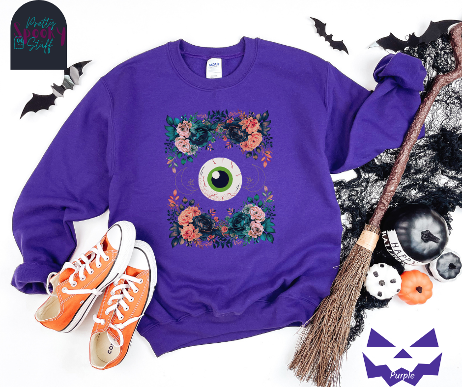 Floral Halloween sweatshirt with a large eyeball in the middle, gold crescent moon and star line drawings on either side, surrounded by four bunches of flowers that make a frame, in purple.