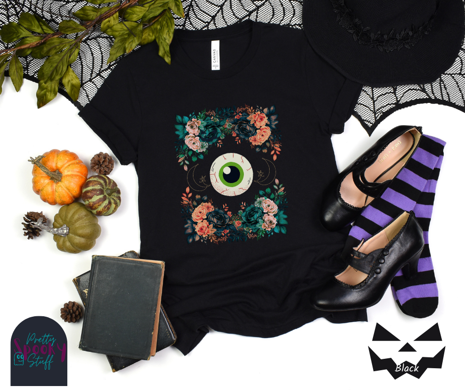Unique Halloween shirt with large eye in the middle, with gold line drawing of crescent moon and star, and four bunches of pink and blue flowers forming a frame around it, in black.