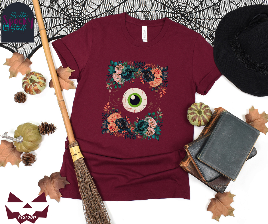 Unique Halloween shirt with large eye in the middle, with gold line drawing of crescent moon and star, and four bunches of pink and blue flowers forming a frame around it, in maroon.