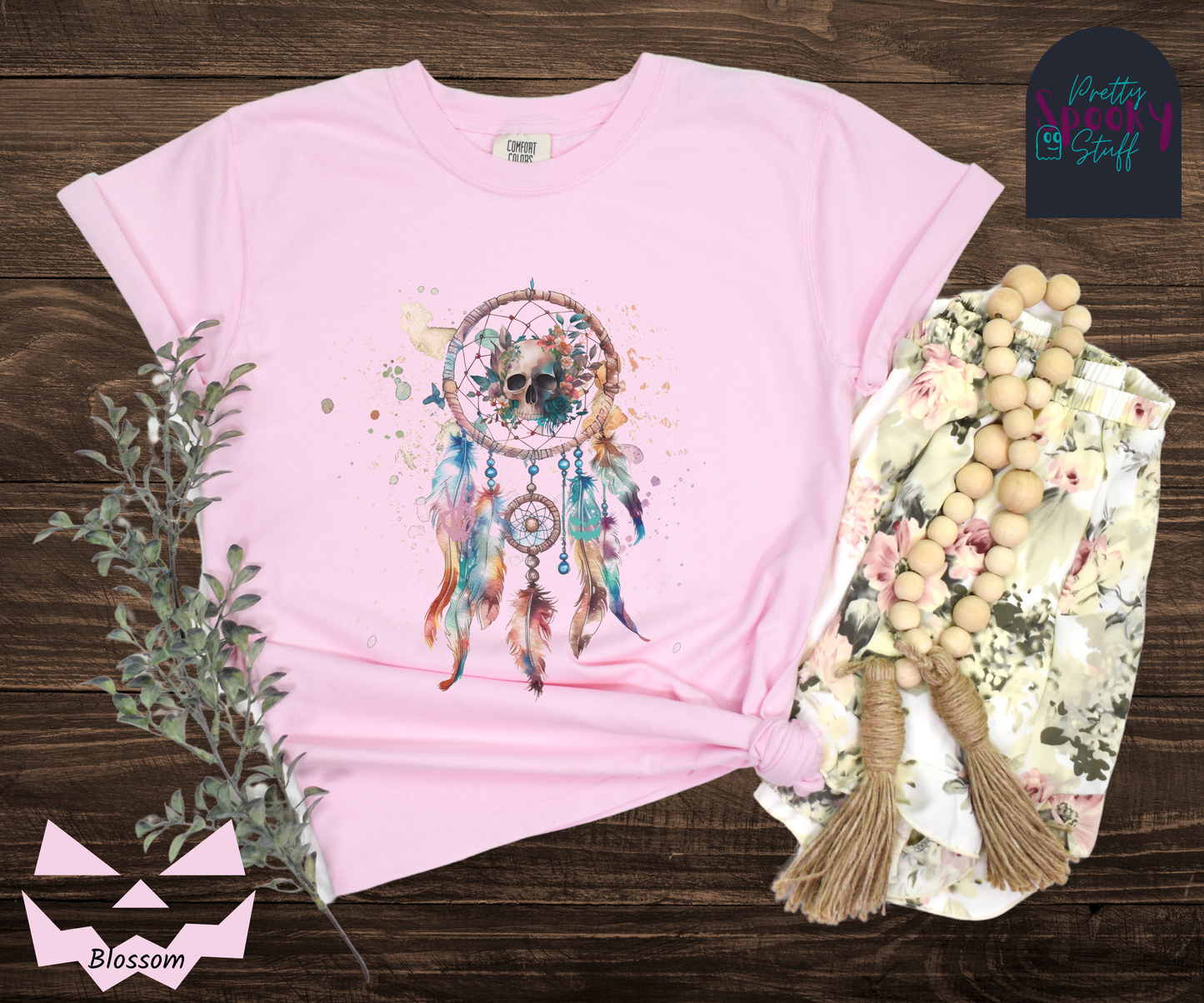 Halloween skull dream catcher shirt, watercolor aesthetic in pinks, blues, and purples, featuring a creepy skull with flowers behind it in the main circle, and skeleton hand and jack o lantern face hidden amongst the 5 feathers hanging below, in blossom.