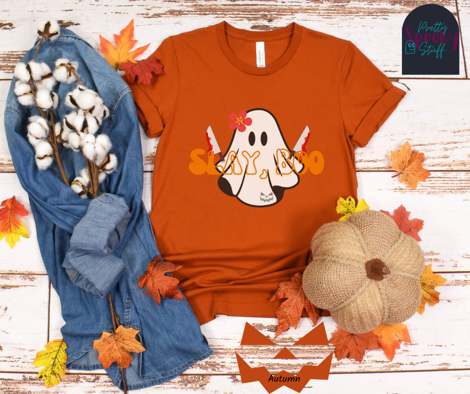 Ghost Halloween shirt with a ghost wearing pink flower, holding two bloody butcher knives and the words "slay boo" in orangle font in front of her, in autumn.