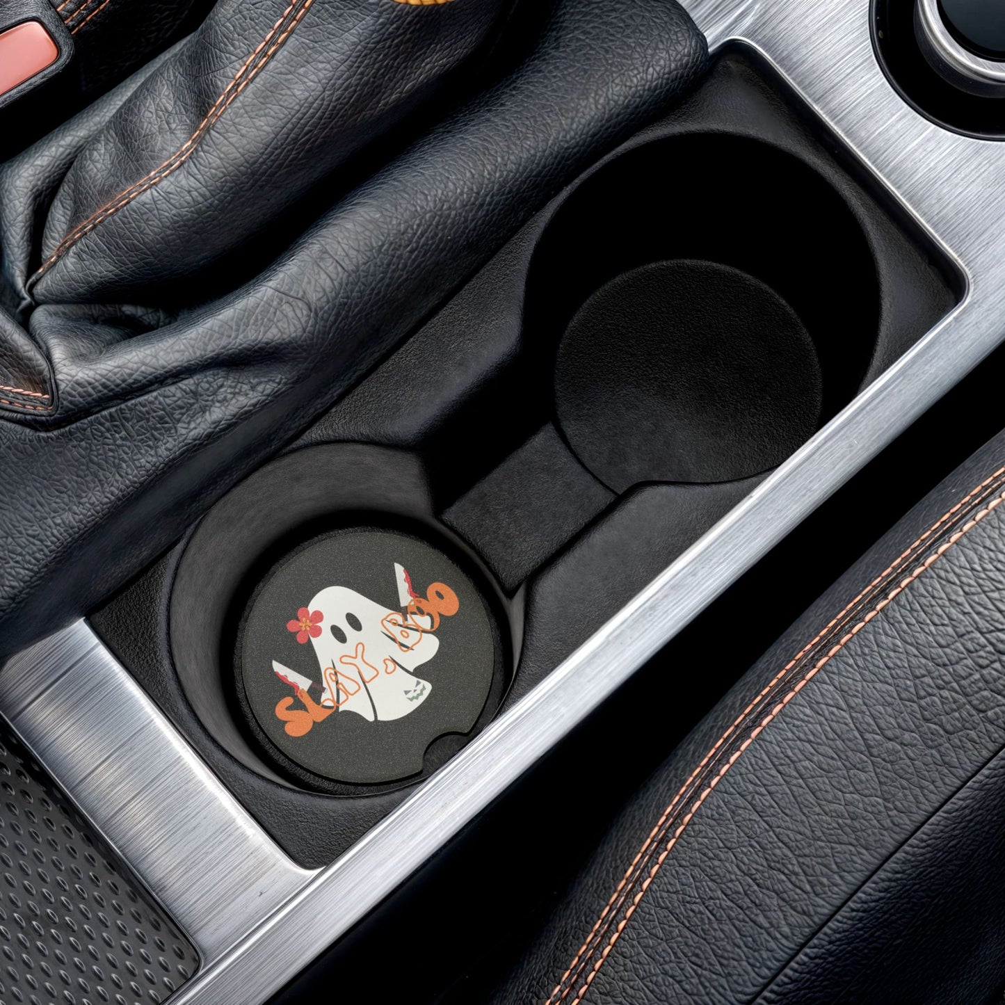 Halloween car coaster with a ghost wearing a pink flower, holding two bloody knives with the words "slay boo" in orange vintage font in front, black background, shown in car cup holder.