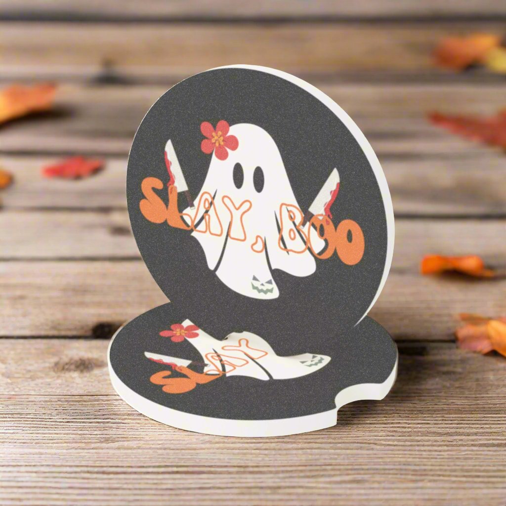 Halloween car coaster with a ghost wearing a pink flower, holding two bloody knives with the words "slay boo" in orange vintage font in front, black background, on table.