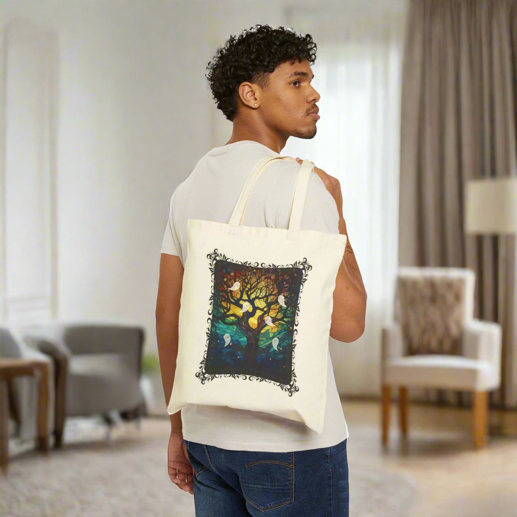 Halloween canvas tote bag with stained glass design featuring a bare brown tree reaching to the glowing moon with ghosts flying all around, natural background, being held over the shoulder of a man.