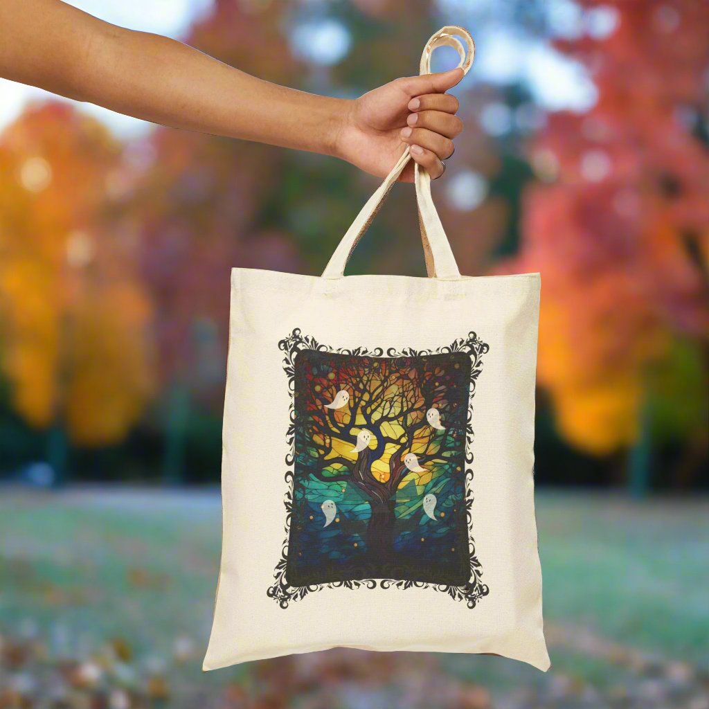 Halloween canvas tote bag with stained glass design featuring a bare brown tree reaching to the glowing moon with ghosts flying all around, natural background, held up by arm.